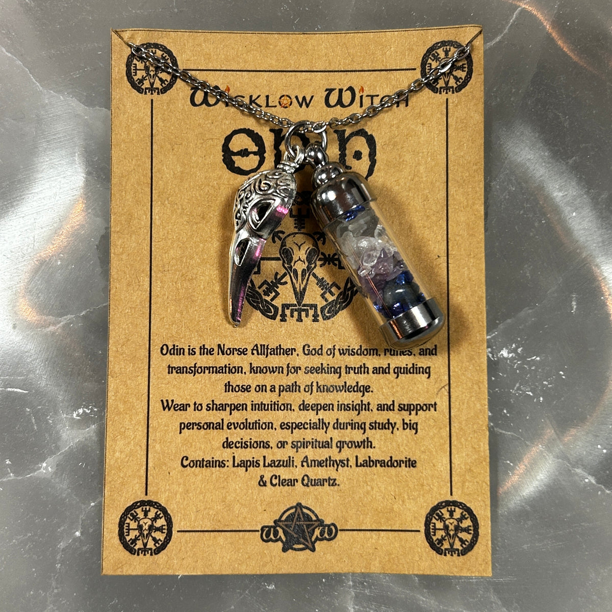 A silver-toned necklace with a raven pendant and a small clear vial pendant filled with purple and blue beads, displayed on a kraft paper backing card labeled 'Wicklow Witch Odin'.