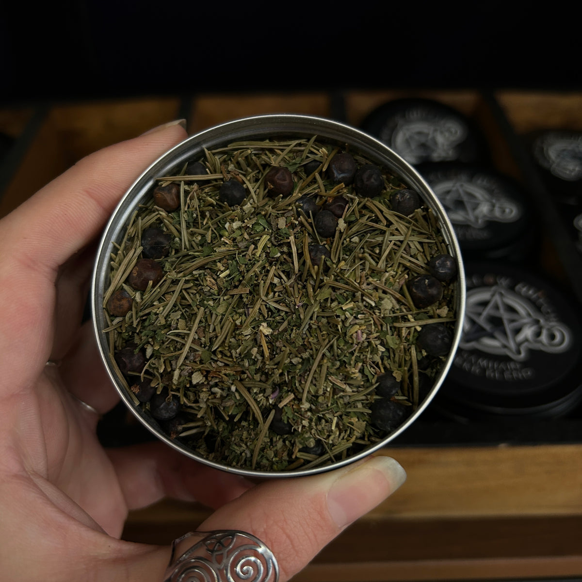 A hand holds a round metal tin filled with dried botanical herbs and dark berries, with black circular tins featuring pentagram designs in the background on a wooden surface.