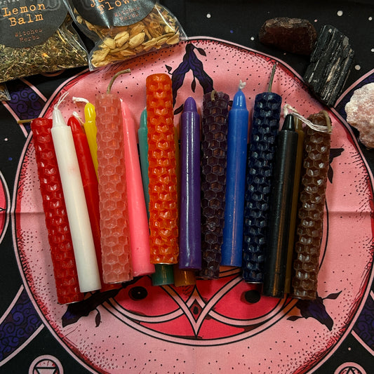 A set of twelve textured beeswax taper candles in various colors, arranged on a pink and black decorative plate. The candles include red, white, orange, pink, green, purple, blue, black, and brown.