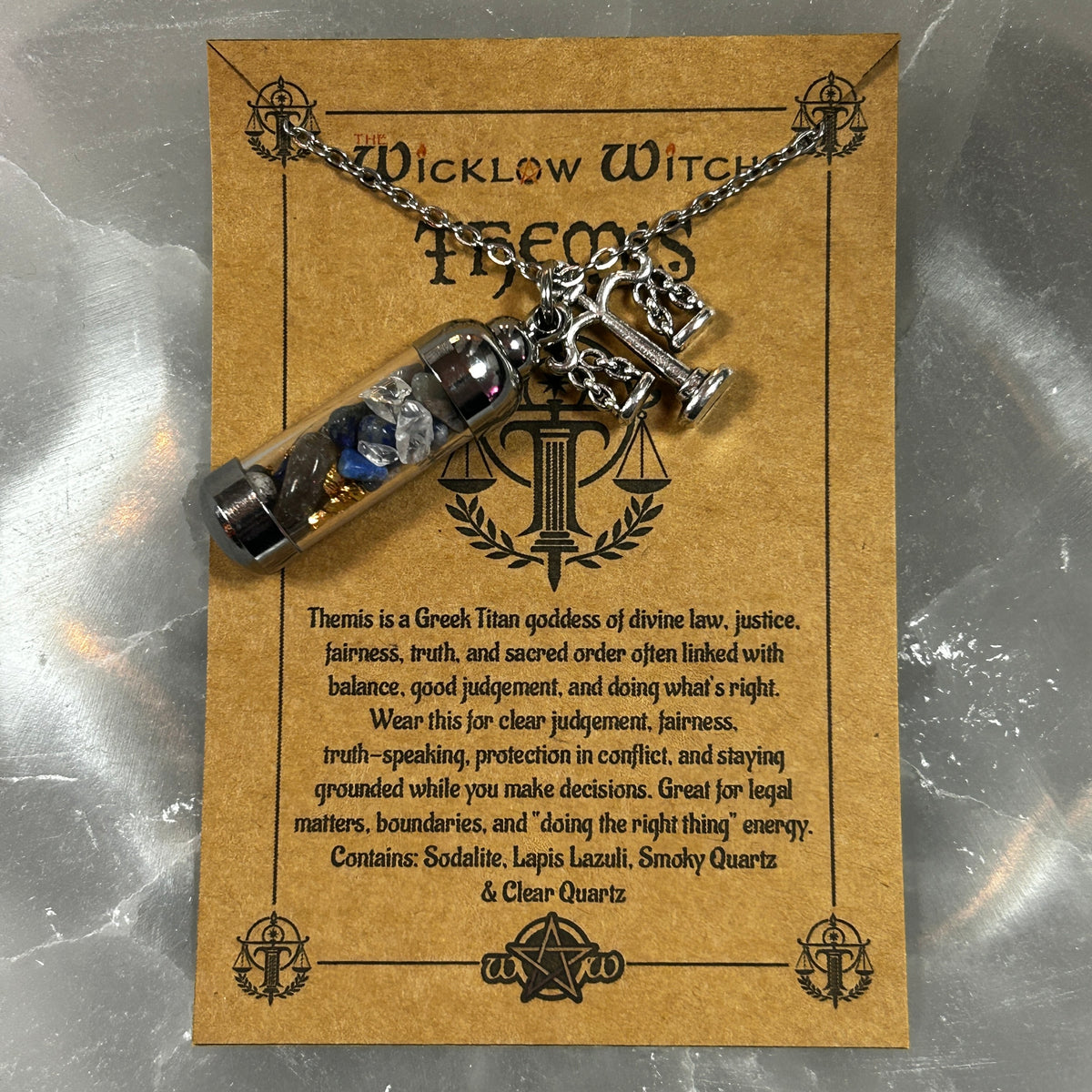 A Wicklow Witch themed pendant necklace featuring a glass vial with blue and silver contents and a silver balance scale charm, displayed on a brown kraft card with text about Themis.