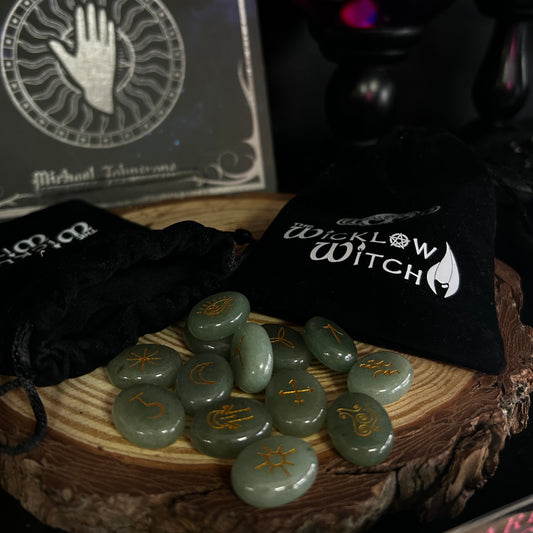 A set of green polished gemstones with gold celestial symbols arranged on a wood slice, alongside black drawstring pouches printed with 'The Willow Witch' logo, with a dark tarot card book and crystal ball in the background.